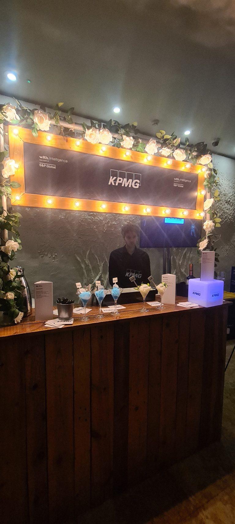 KPMG branded rustic wooden mobile bar with illuminated marquee bulb signage, floral garland, and professional bartender serving corporate cocktails