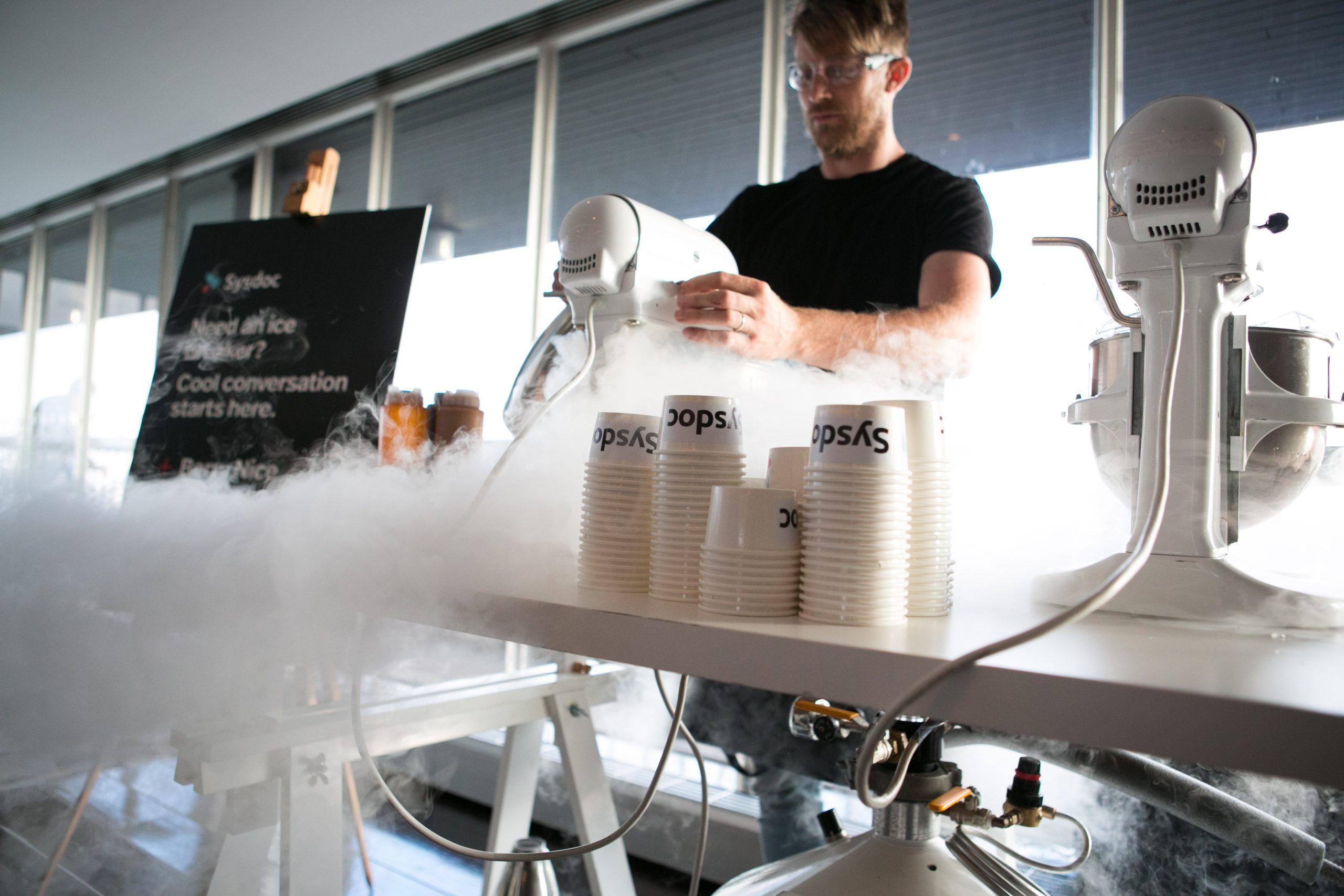 Chef operating stand mixers with dense liquid nitrogen fog cascading over counter with Sysdoc branded cups at corporate event