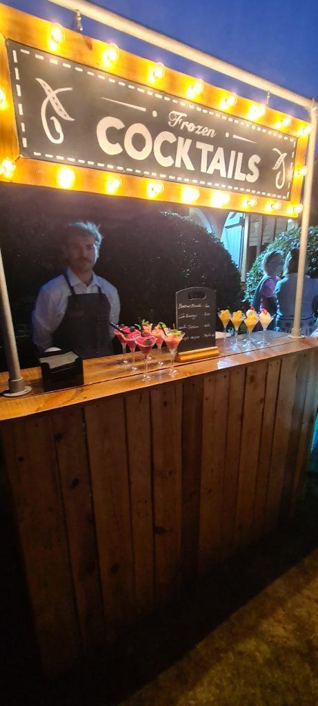 Evening scene of a bartender behind rustic timber bar with warm illuminated marquee bulb sign in outdoor wedding venue