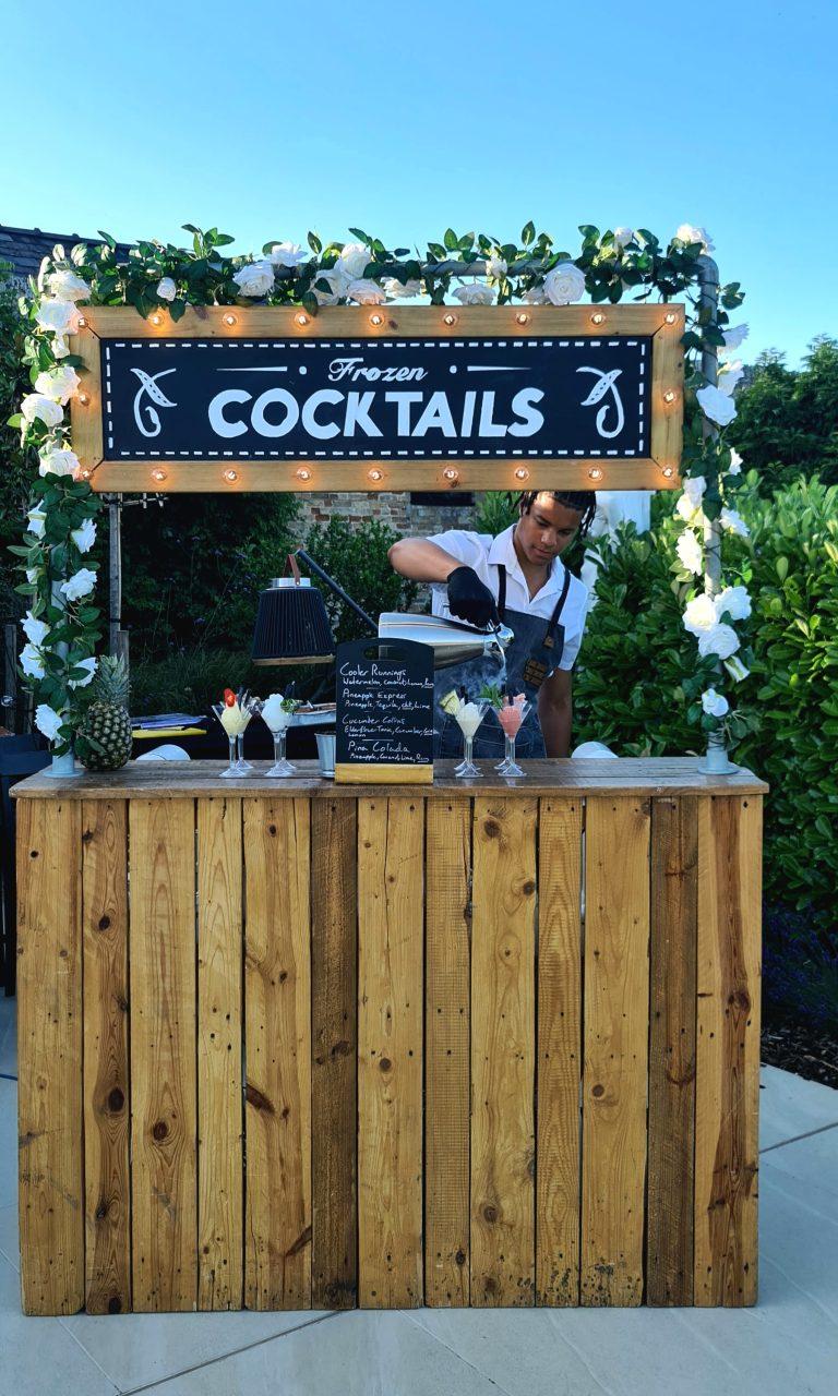 Rustic wooden mobile cocktail bar for wedding with illuminated marquee sign, floral garland, and mixologist crafting frozen drinks